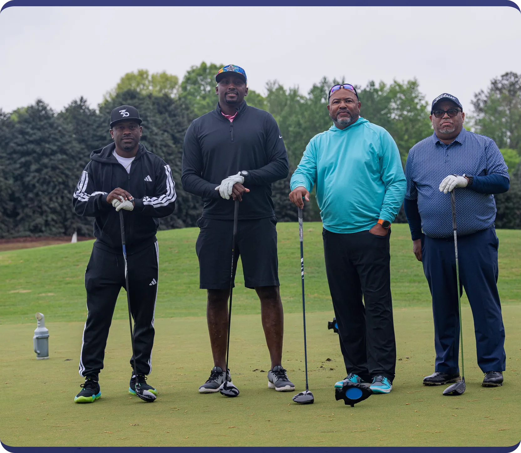 Four men standing on a golf course holding golf clubs, wearing casual sportswear and golf gloves.