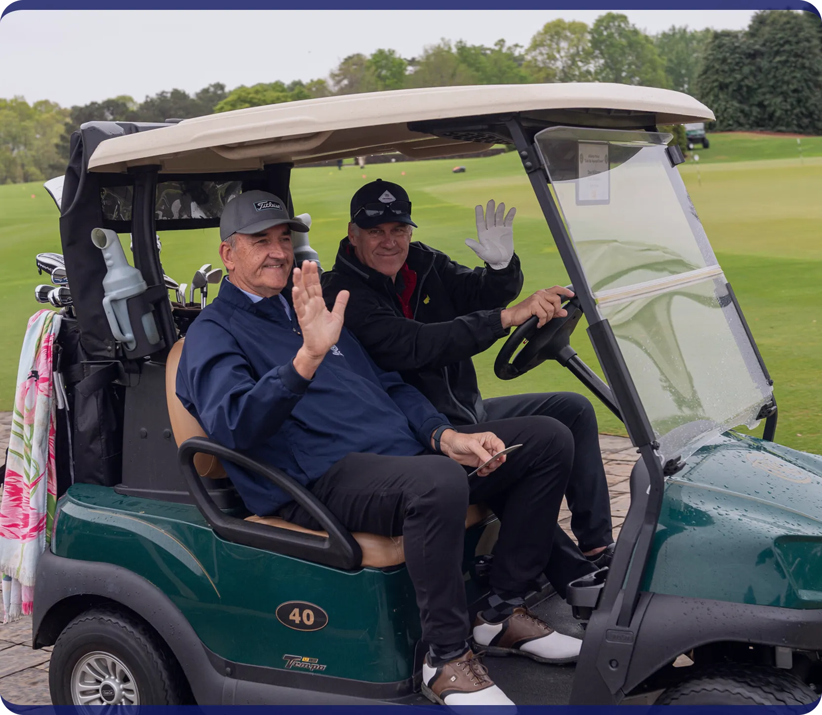 Two men sitting in a green golf cart on a golf course waving at the camera.