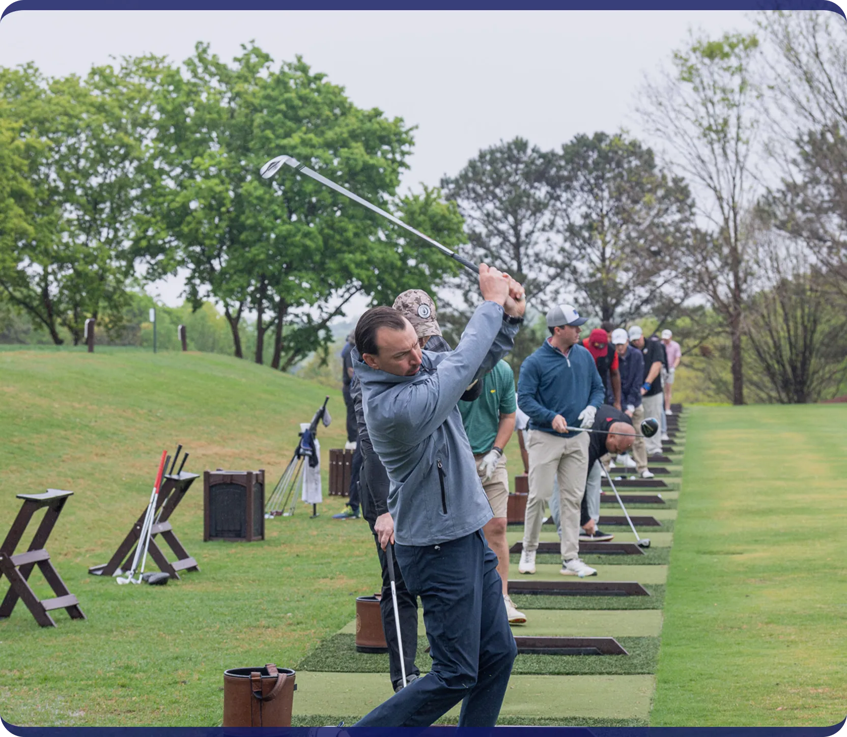 Golfers practicing their swings in a row at a driving range with green grass and trees in the background.