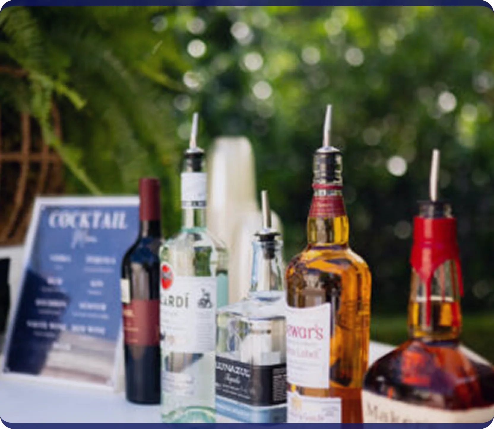 Five liquor bottles with pour spouts on a table outdoors next to a cocktail menu sign.