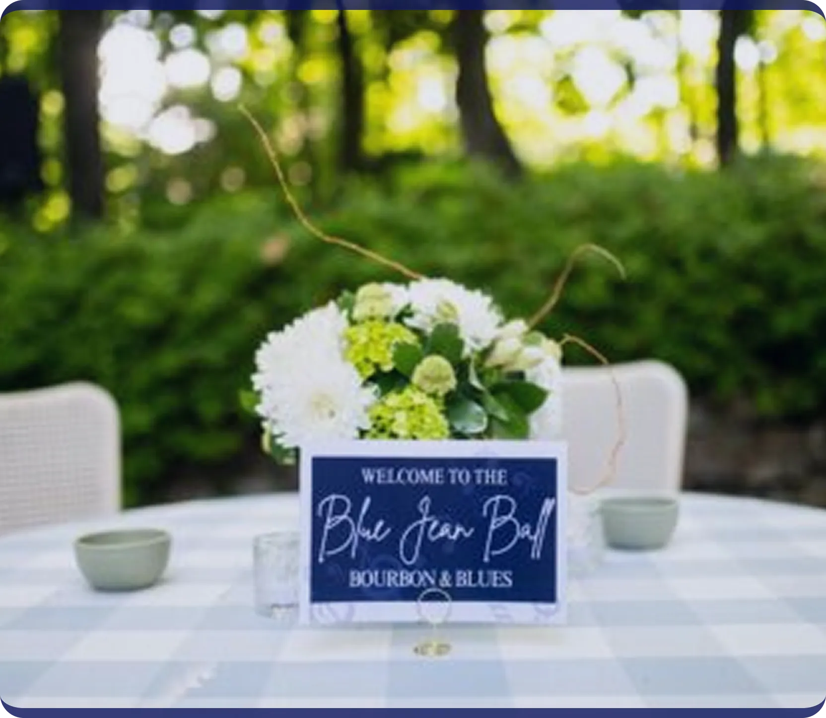 Table with a white checkered tablecloth, a flower centerpiece, and a sign reading 'Welcome to the Blue Jean Ball Bourbon & Blues' outdoors.