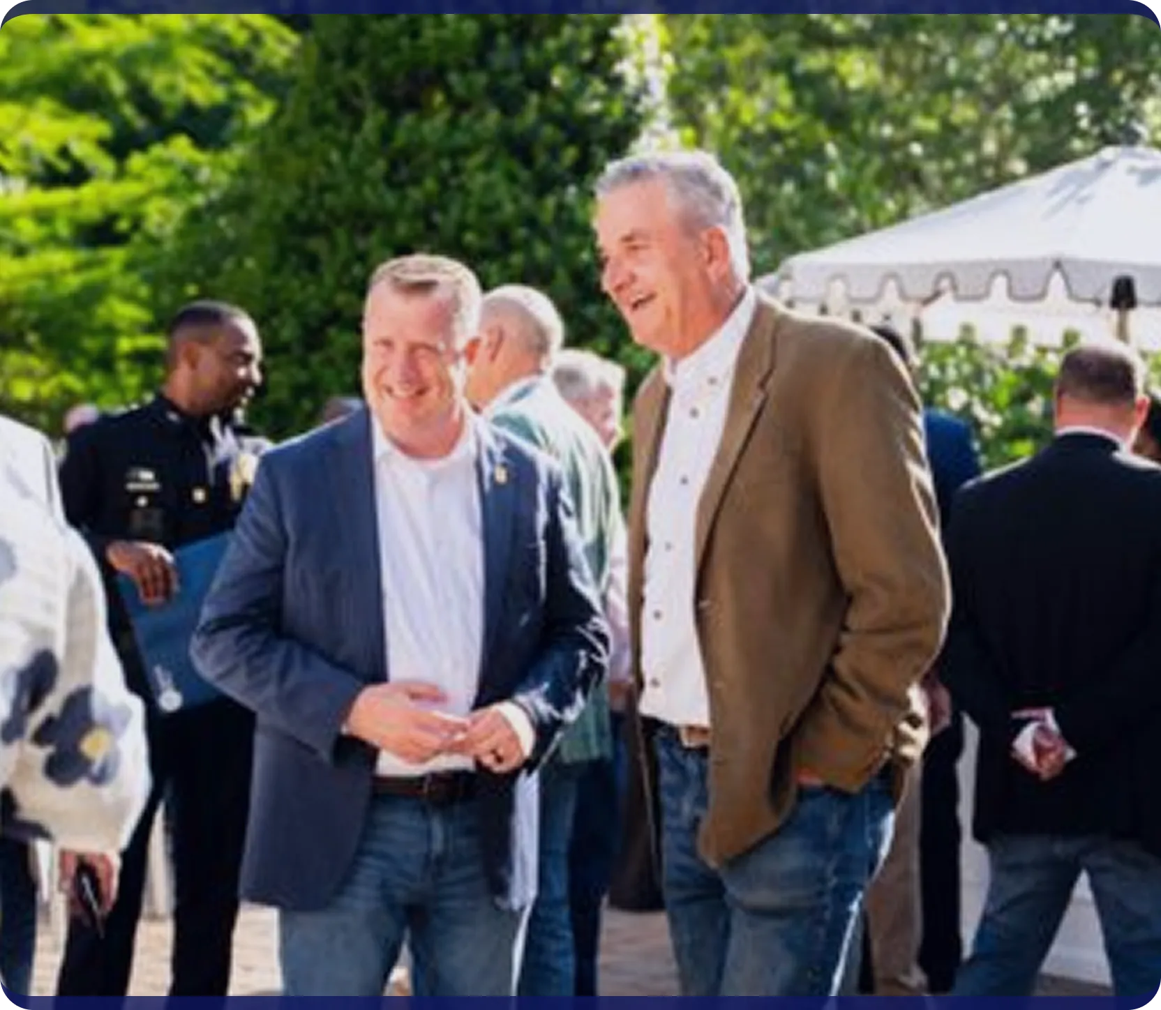 Two middle-aged men smiling and chatting outdoors at a social gathering with others in the background.