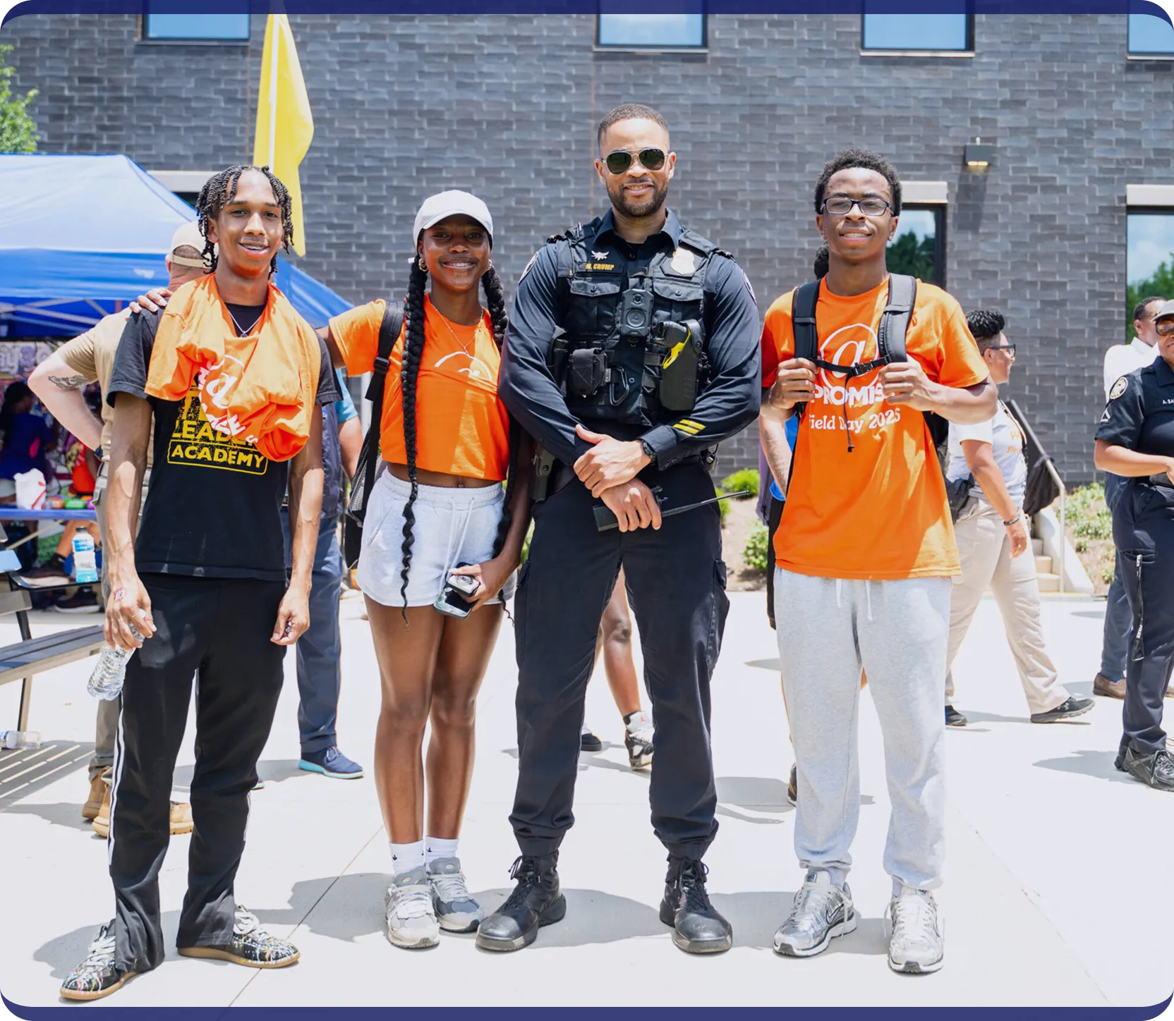 Three young people wearing orange Promise Academy shirts pose with a uniformed police officer outside on a sunny day.