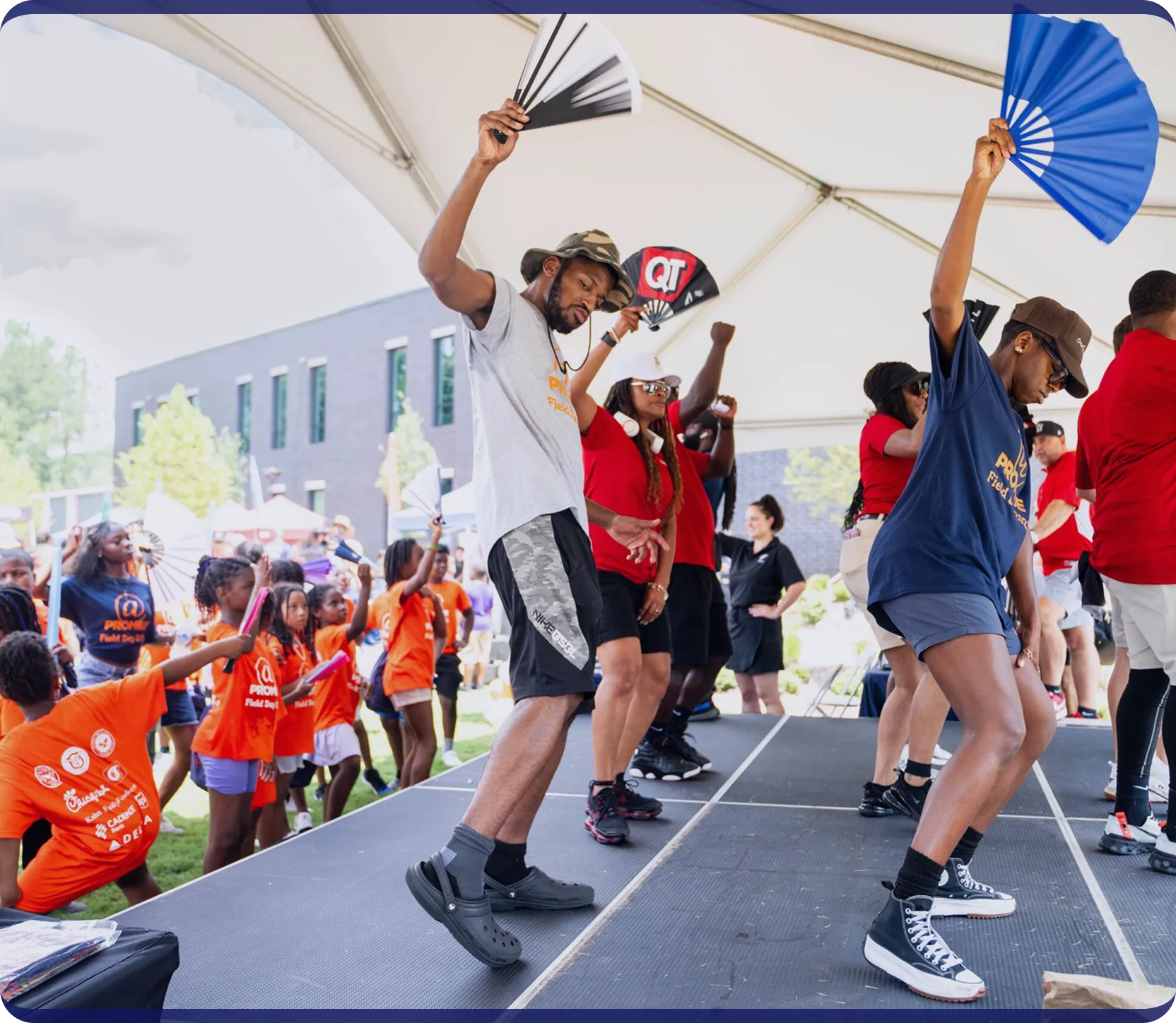 Group of people dancing on a stage under a white canopy, some holding fans, with children in orange shirts dancing and watching nearby outdoors.