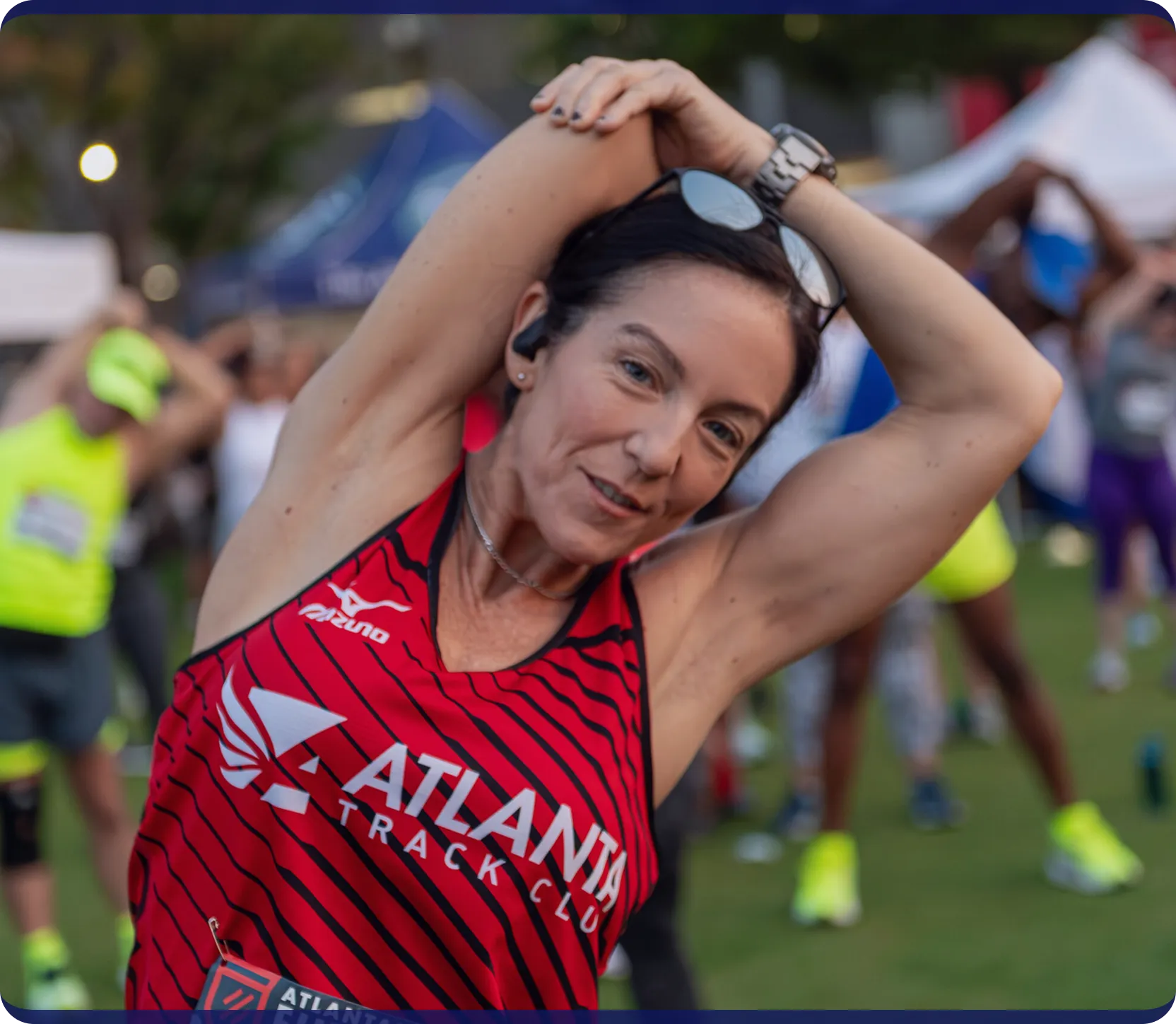 Woman in an Atlanta Track Club shirt stretching her arm over her head at an outdoor event.