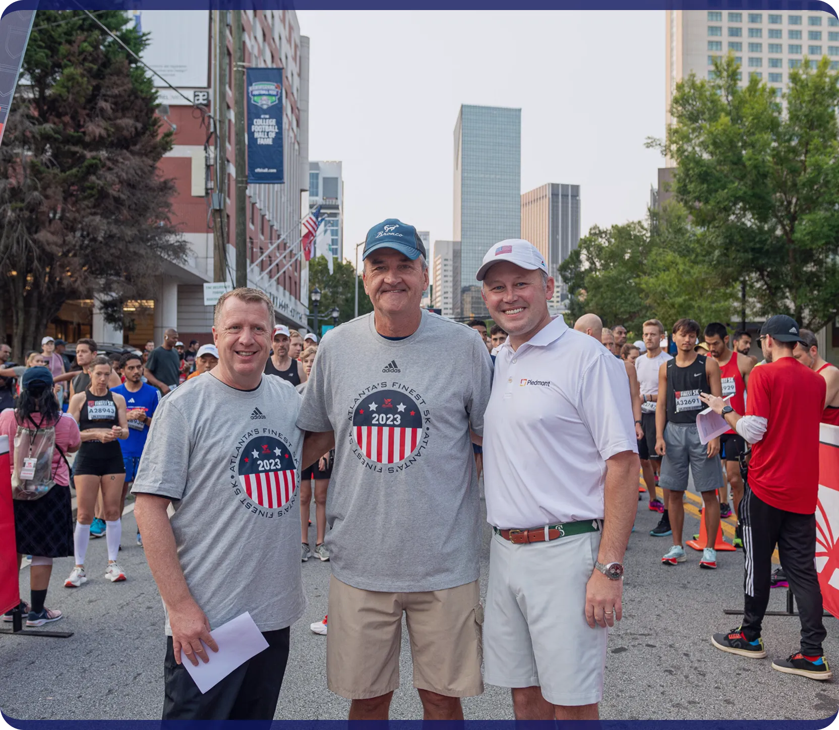 Three men smiling at a starting line of a 2023 Atlanta's Finest 5K race with runners in the background.