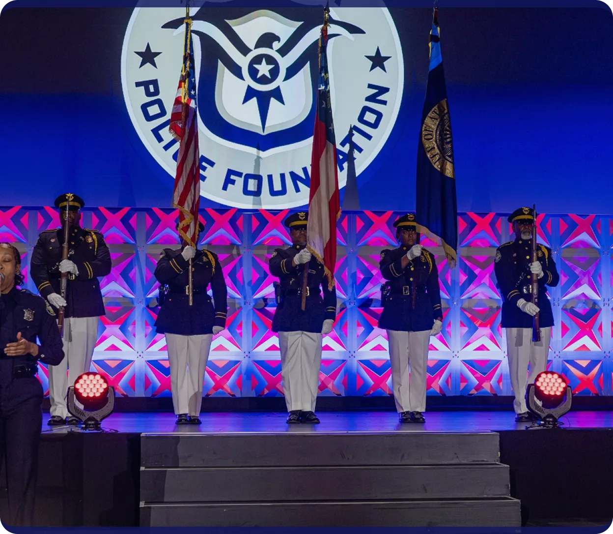 Police honor guard in formal uniforms holding flags on stage with a Police Foundation emblem projected in the background.