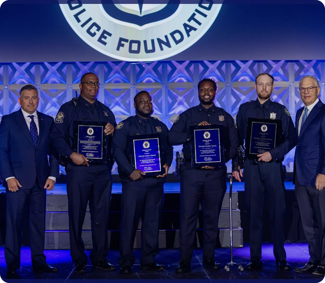 Five police officers holding plaques posing for a photo with two men in suits under a Police Foundation sign.