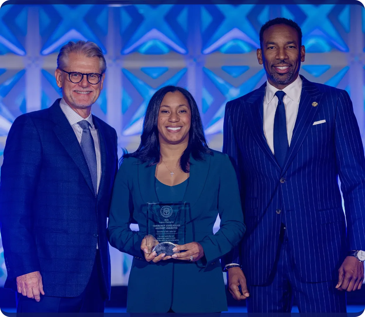 Three professionally dressed people smiling, with the woman in the center holding an award.