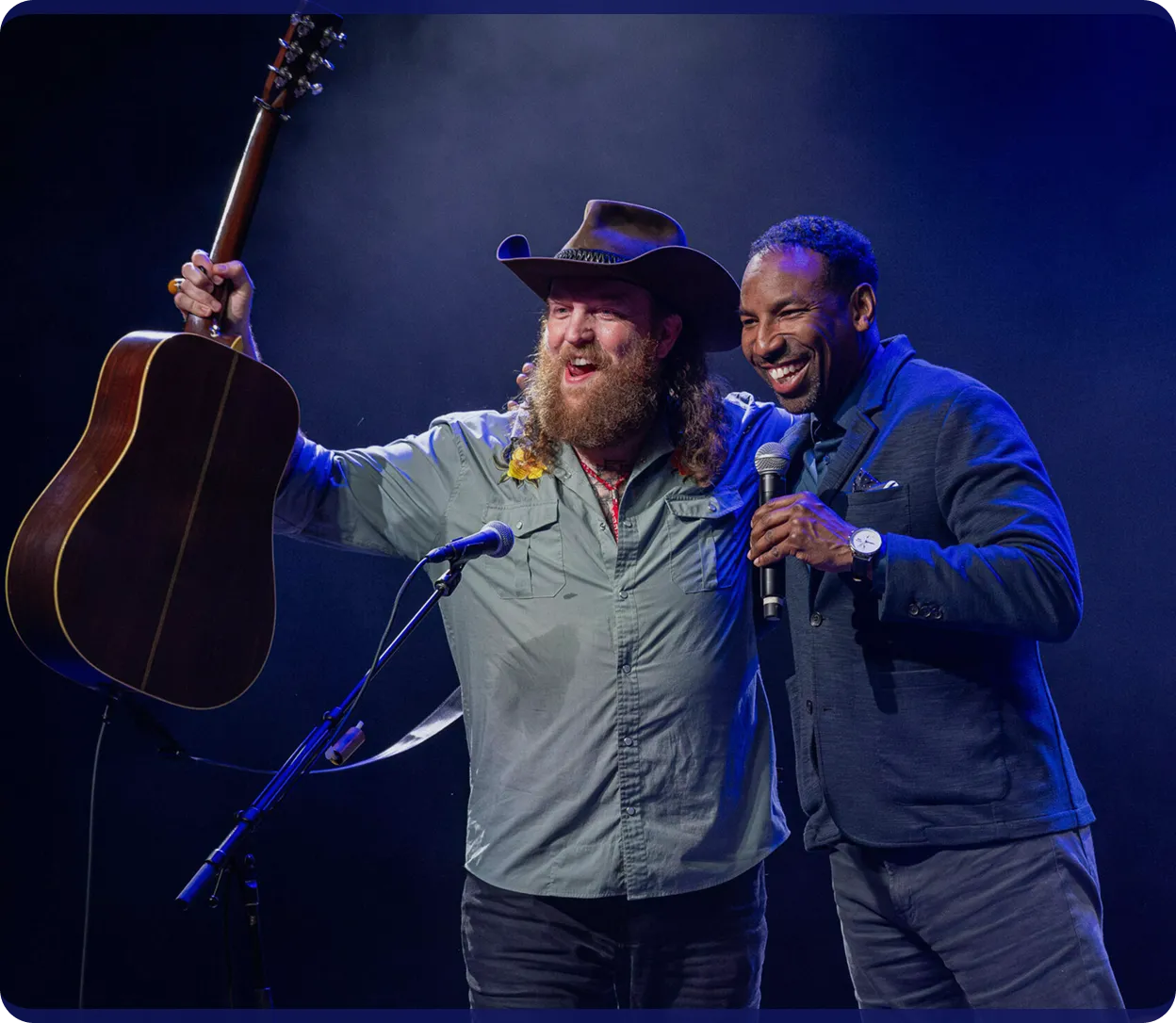 Two men on stage smiling, one holding a guitar up and the other holding a microphone.