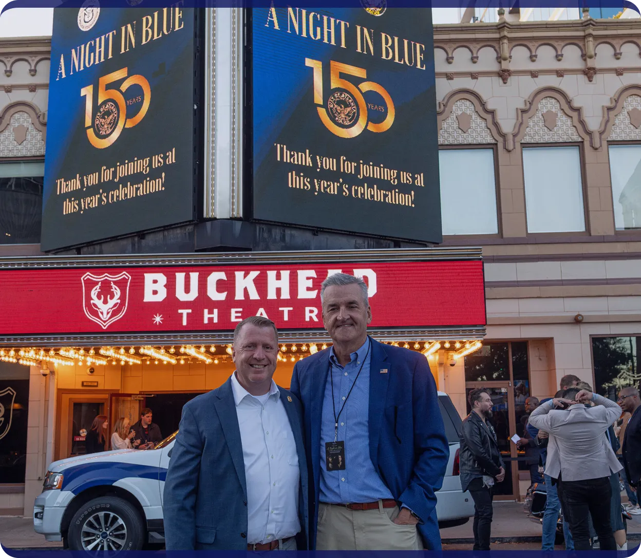 Two men in suits posing and smiling in front of Buckhead Theatre with event signage reading 'A Night in Blue 150 Years'.