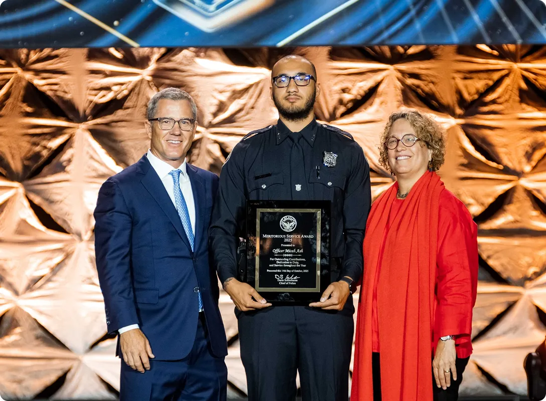 Police officer in uniform holding a Meritorious Service Award plaque, standing between a man in a blue suit and a woman in a red outfit, all smiling.