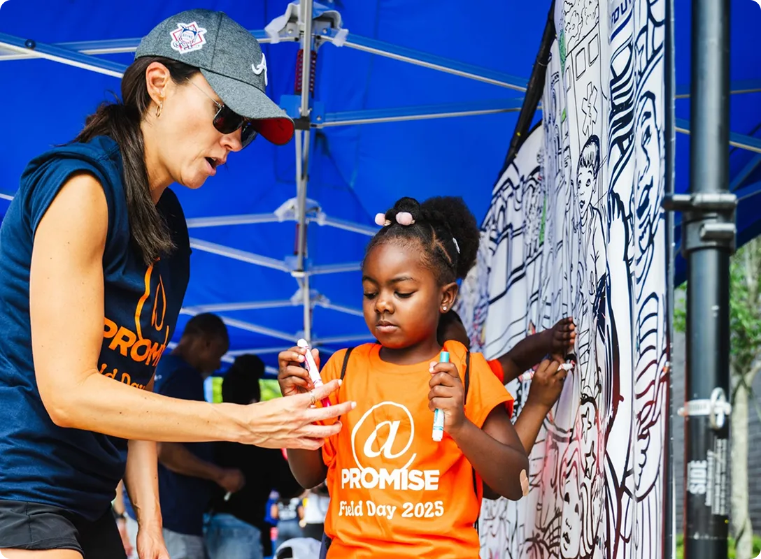 A young girl in an orange 'Promise Field Day 2025' shirt holds markers while a woman wearing a cap and sunglasses helps her under a blue event tent.