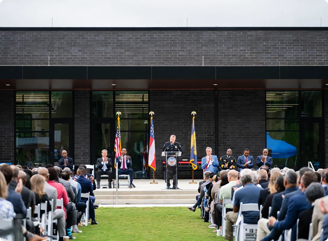 Police officer speaking at a podium on an outdoor stage with American and state flags, surrounded by seated officials and an audience.