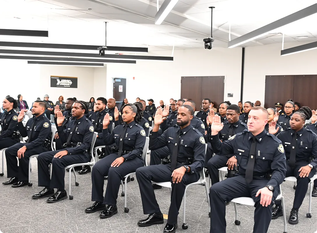 A group of uniformed police officers sitting in rows indoors, raising their right hands as if taking an oath.