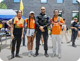 Three young people wearing orange shirts standing with a smiling police officer on a city street.