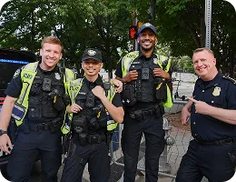 Four smiling police officers, three wearing neon yellow safety vests and one in a black uniform, standing outdoors with trees and a parked police vehicle behind them.