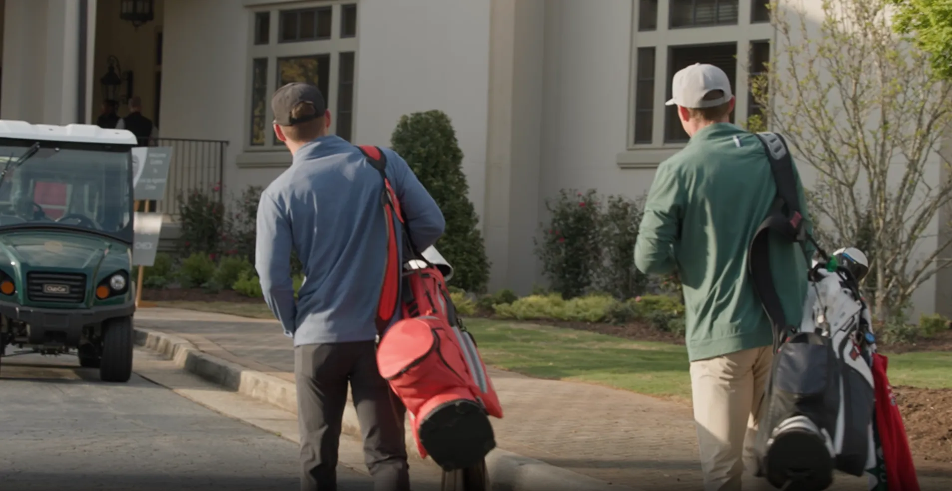Two men carrying golf bags walking on a path near a golf cart and a building.