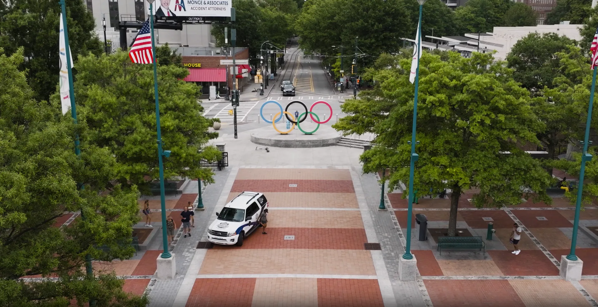 Urban plaza with trees, an American flag, police vehicle, pedestrians, and large Olympic rings sculpture in the center.