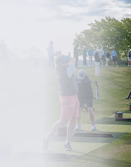 Golfers practicing swings on a driving range under a cloudy sky with a tree and group of people in the background.