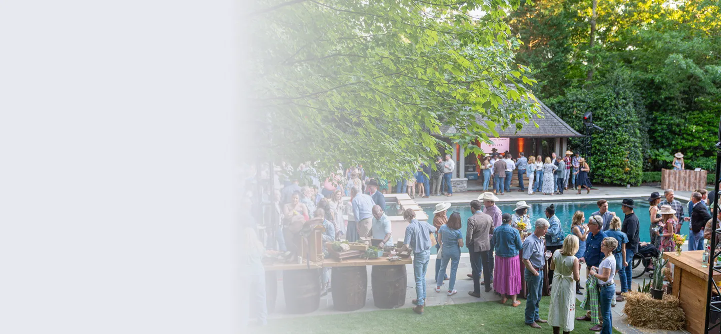 Outdoor party with people socializing around a pool, some wearing hats, under trees near a wooden cabin.