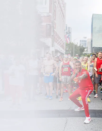 A man in red athletic wear poses energetically in front of a group of runners at the start of a race in an urban setting.