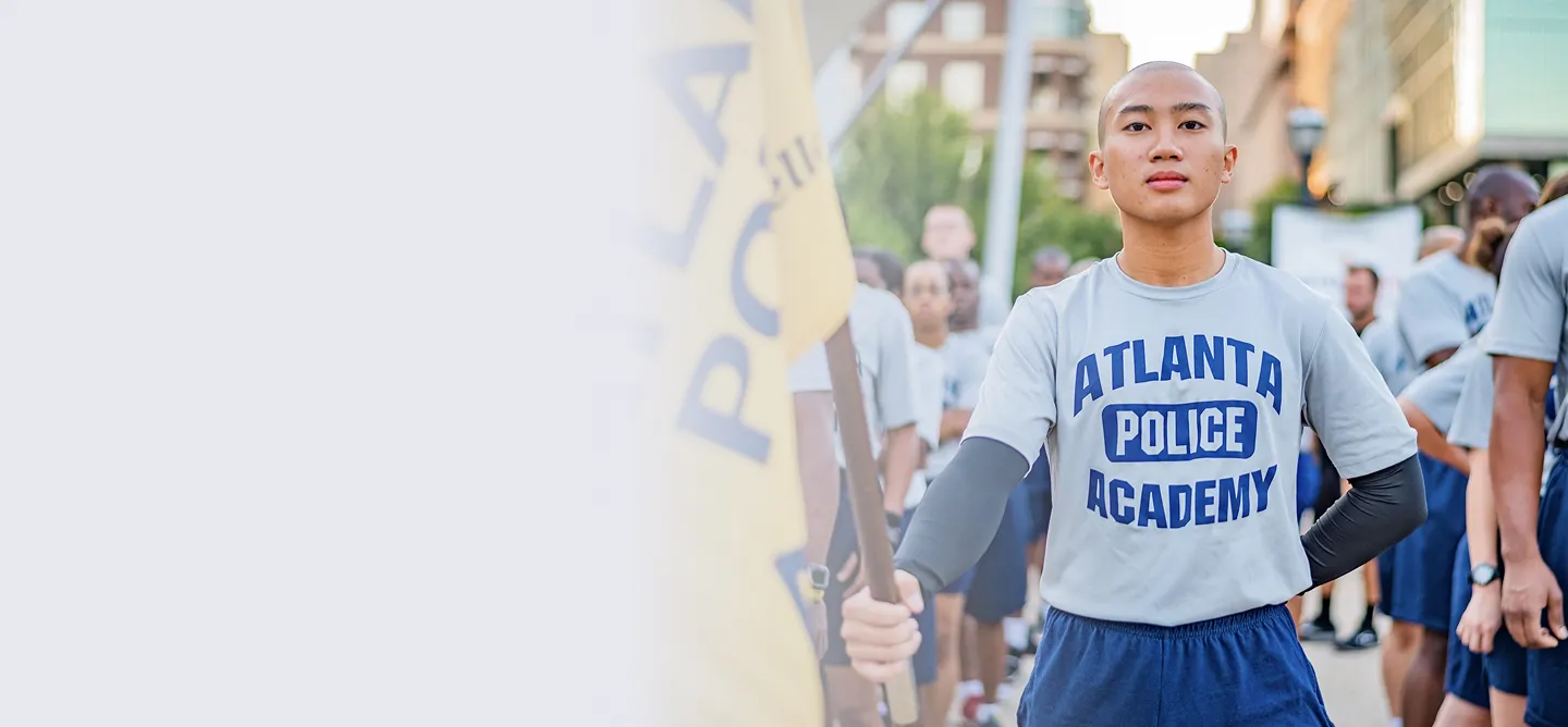 Young person standing with one hand on hip and holding a flag, wearing an Atlanta Police Academy t-shirt with a group of people behind.