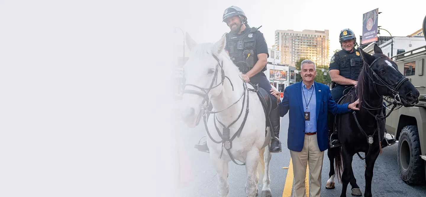 Two police officers on horseback standing beside a man in a blue blazer on a city street at sunset.