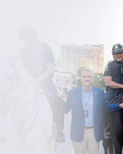 A man in business casual attire standing between two police officers on horseback in an urban setting with a building in the background.
