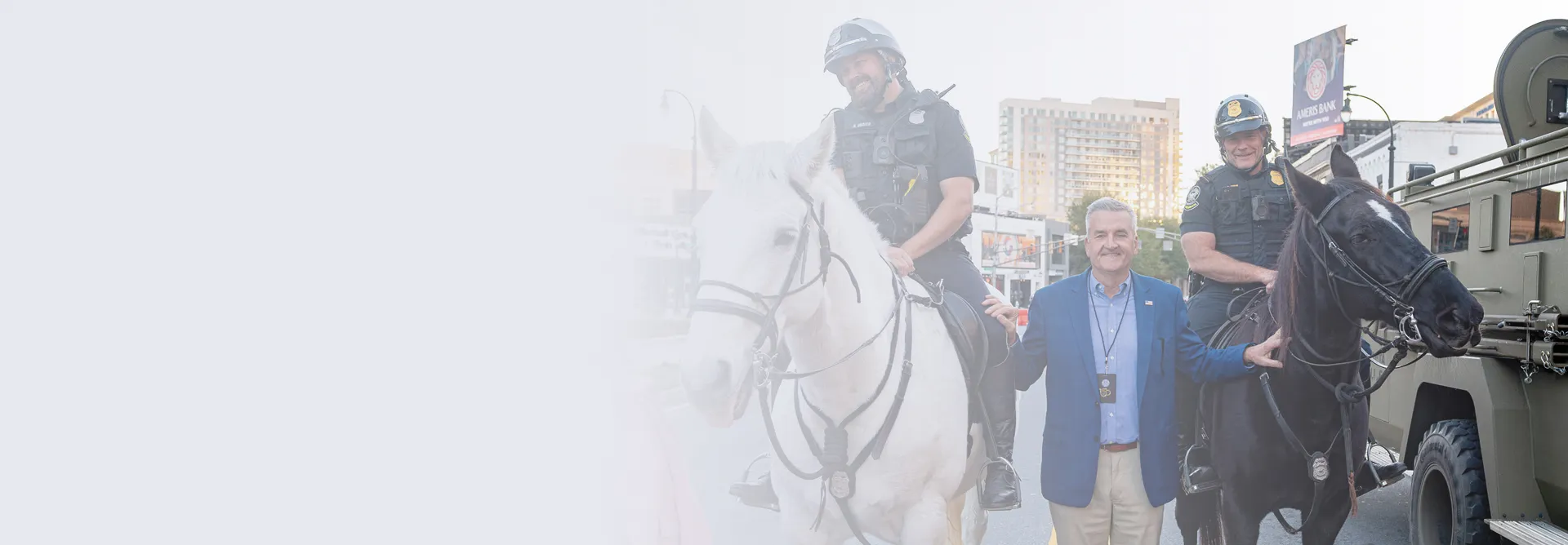 Two mounted police officers on horses beside a smiling man in a blue blazer standing on a city street.