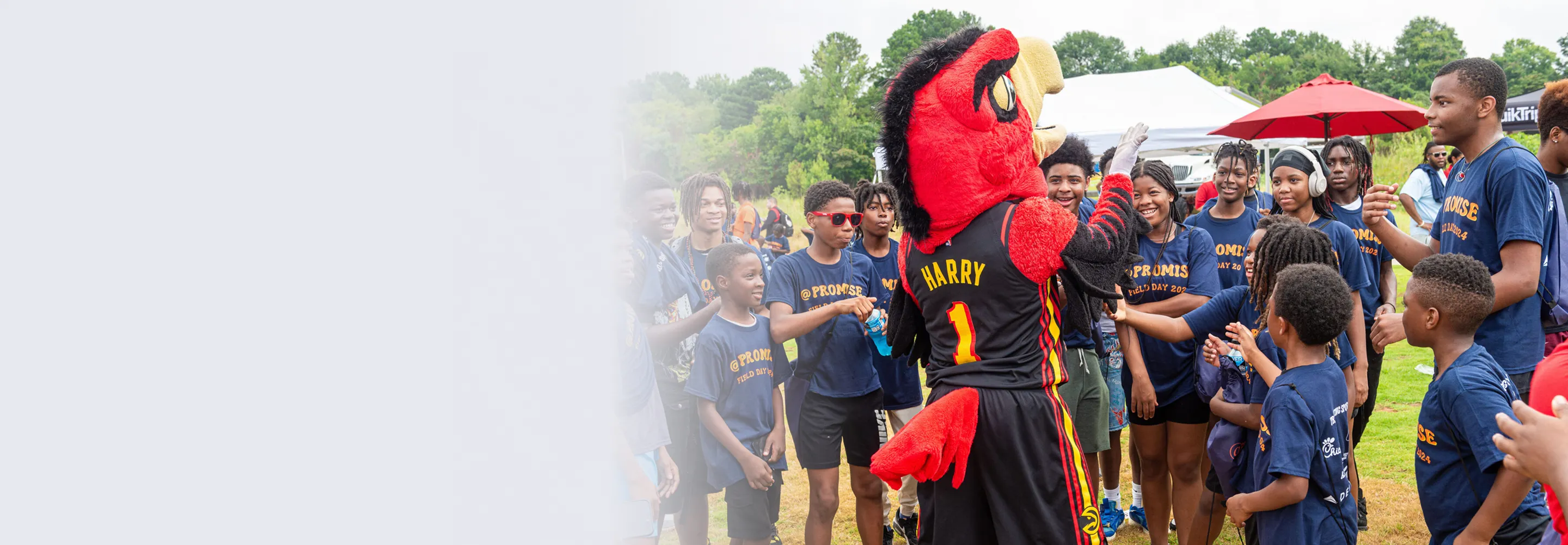 Group of children and teenagers wearing blue @PROMISE field day shirts interacting with a red and black mascot named Harry outdoors.