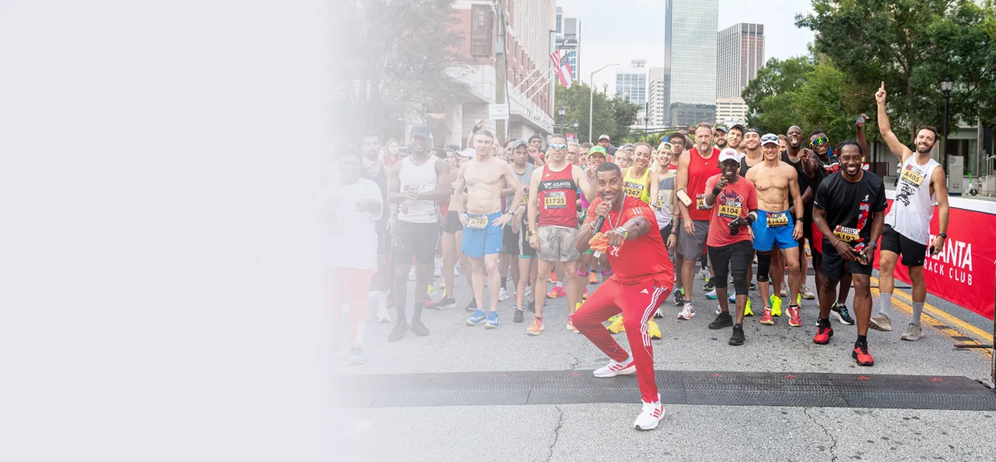 Group of diverse runners gathered at a starting line for a race on a city street with a man in red in front signaling and holding a microphone.