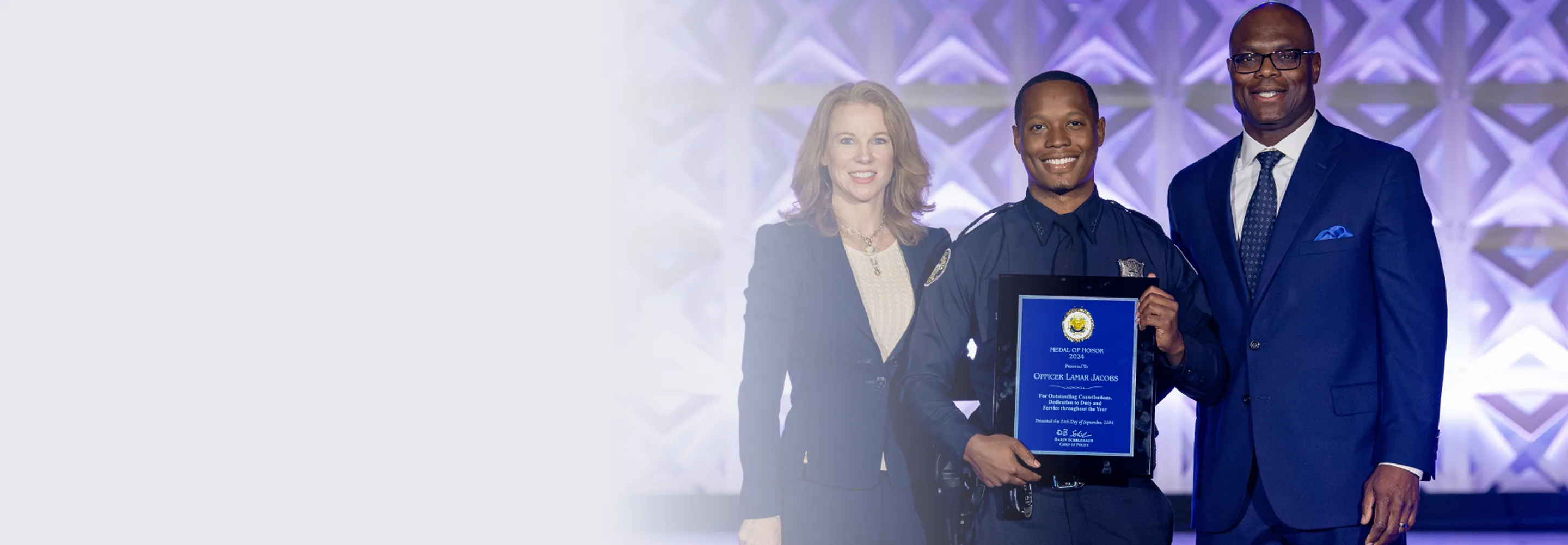 Smiling police officer holding a framed Medal of Honor award, standing between a woman and a man in suits.