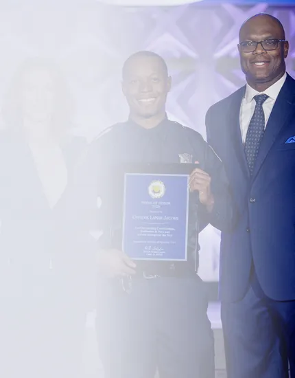 Smiling police officer holding a certificate, standing next to a man in a suit and tie.