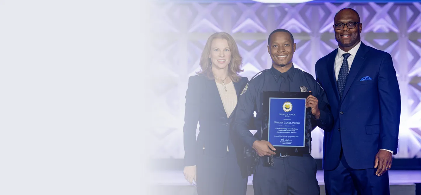 Smiling police officer holding a Medal of Honor plaque, standing between a woman and a man in formal attire on a stage with purple lighting.