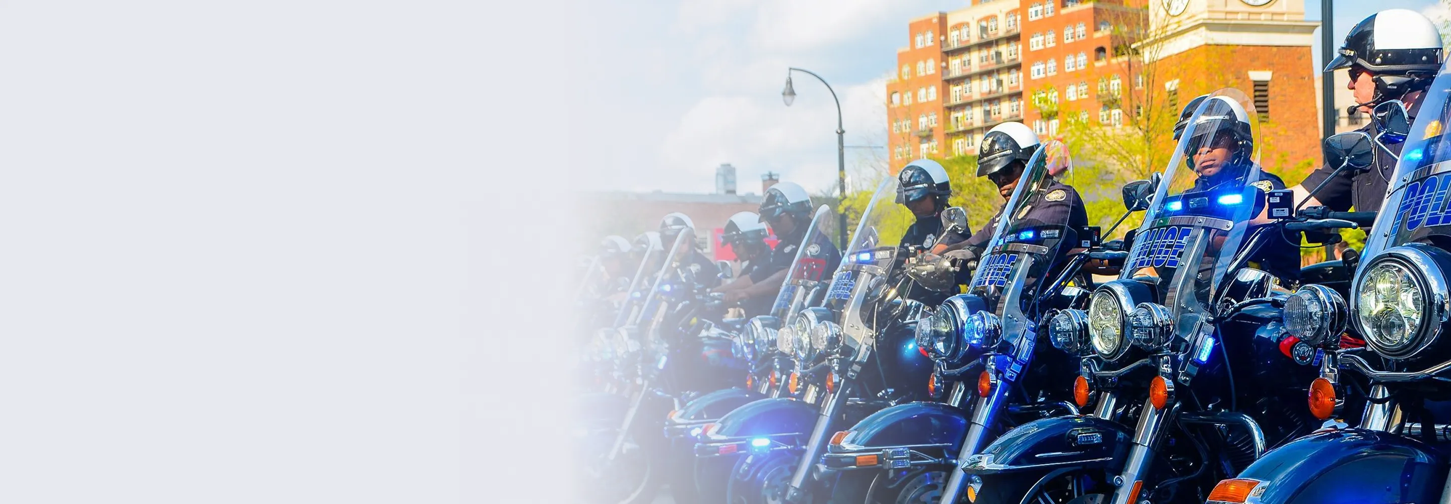 Row of police officers on motorcycles with flashing blue lights parked in an urban area during the day.