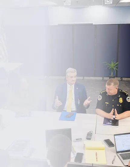 Two men seated at a table in a meeting room, one in a police uniform and the other in a suit, with a laptop, notepad, and phones on the table.