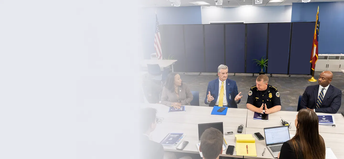 Group of professionals, including a police officer and business people, having a meeting around a conference table in an office room with American flags in the background.
