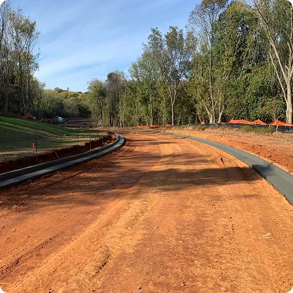 Curved dirt road under construction with concrete curbs on both sides, surrounded by trees and grassy areas under a blue sky.