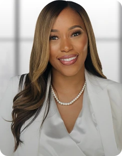 Smiling woman with long, smooth hair wearing a white blouse and pearl necklace against a blurred window background.