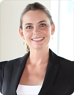 Smiling woman with light brown hair tied back, wearing a black blazer and white top, in a bright room.