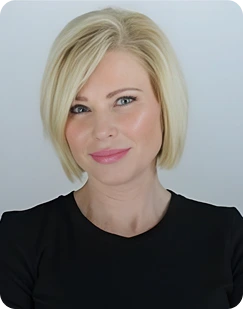 Blonde woman with a short bob hairstyle wearing a black top, smiling softly against a light gray background.