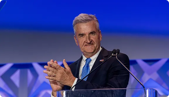 Man in a dark suit and blue tie clapping while standing behind a lectern with microphones, against a blue and gray background.