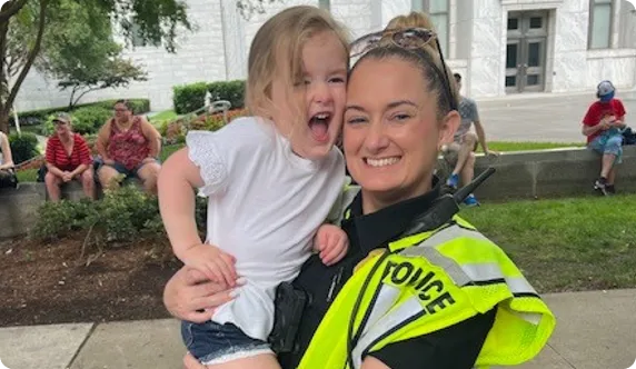 Smiling police officer in a neon yellow vest holding a happy young girl in casual clothes outdoors.