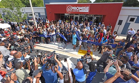 Crowd gathered for a ribbon-cutting ceremony in front of the @PROMISE Center building with many photographers and attendees.