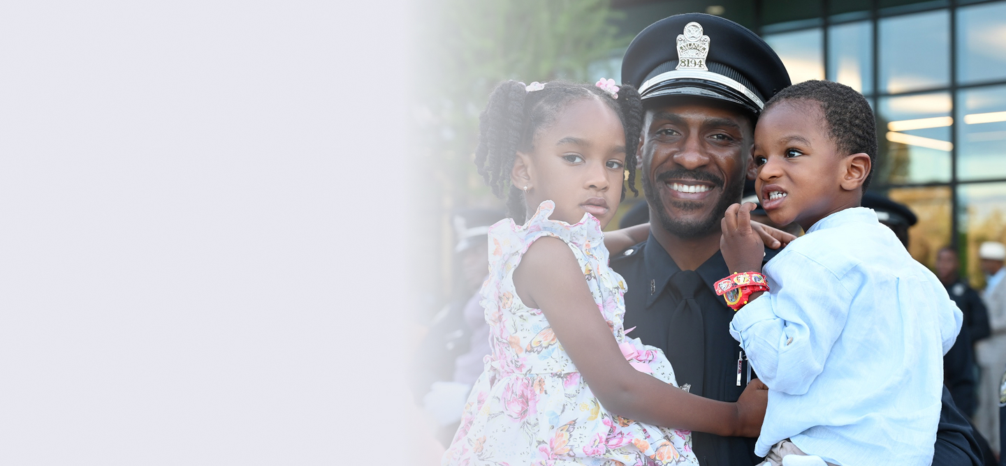 Smiling police officer in uniform holding a young girl in a floral dress and a young boy in a light blue shirt.