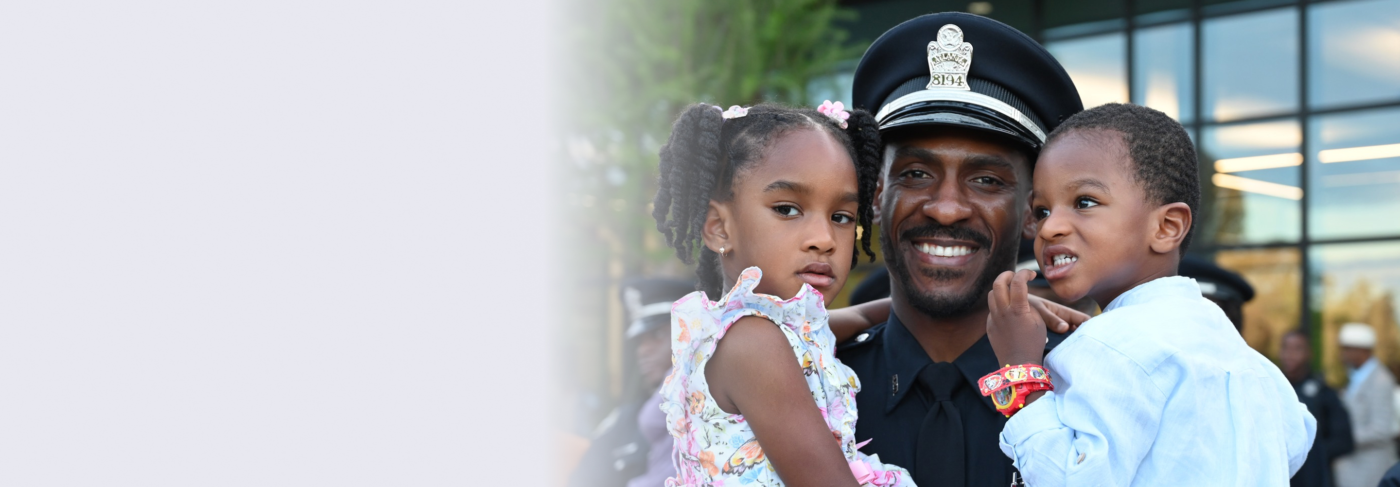 Smiling police officer in uniform holding a young girl and boy outside a building with glass windows.