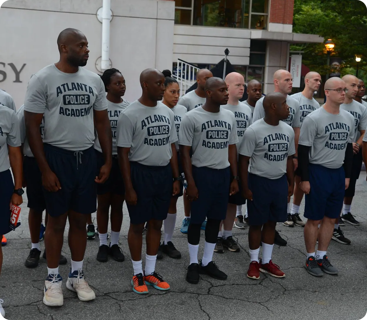 Group of diverse men and women in matching Atlanta Police Academy T-shirts and shorts standing in formation outdoors.