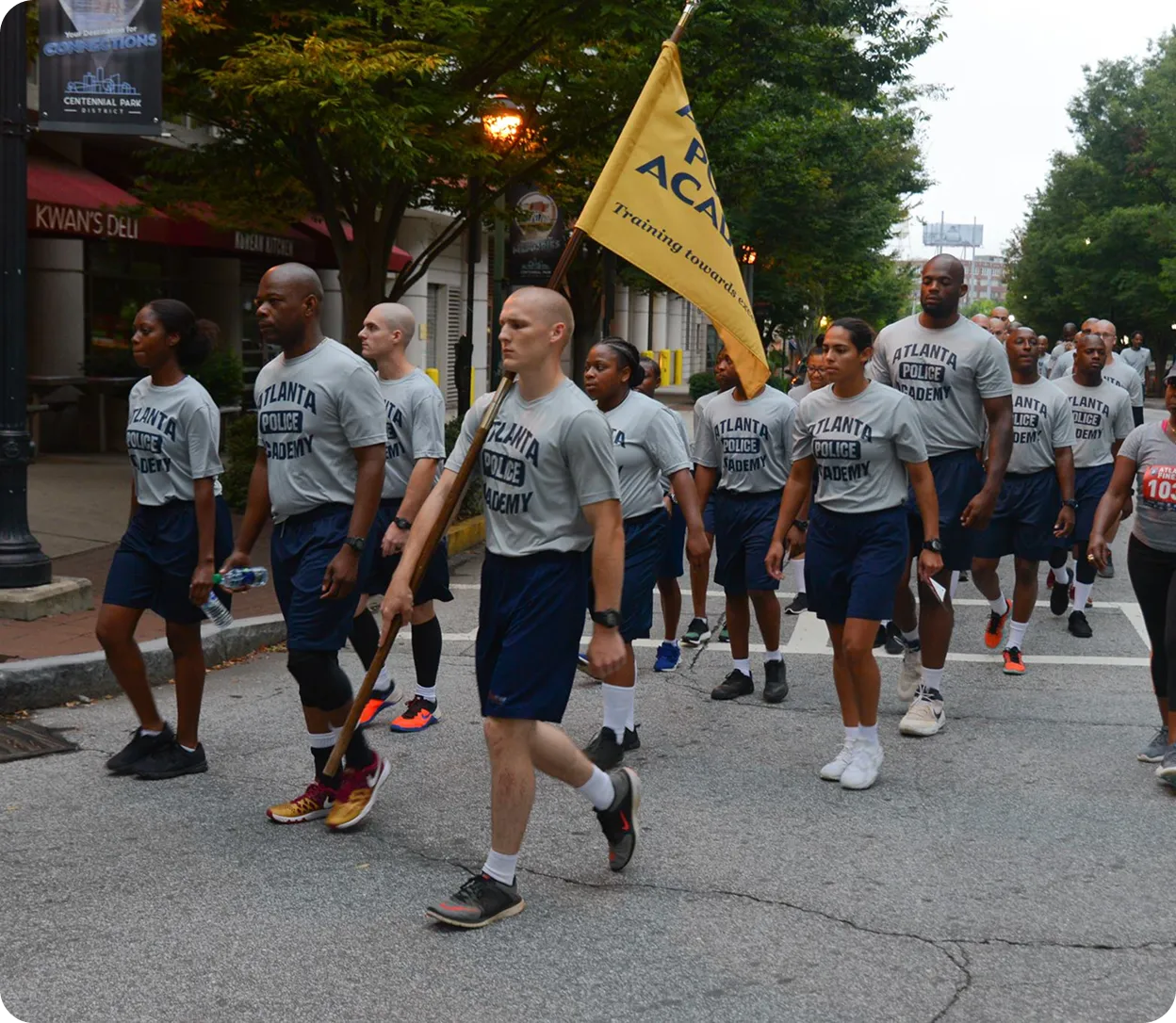 Group of Atlanta Police Academy trainees marching in formation on a city street, one holding a yellow flag.