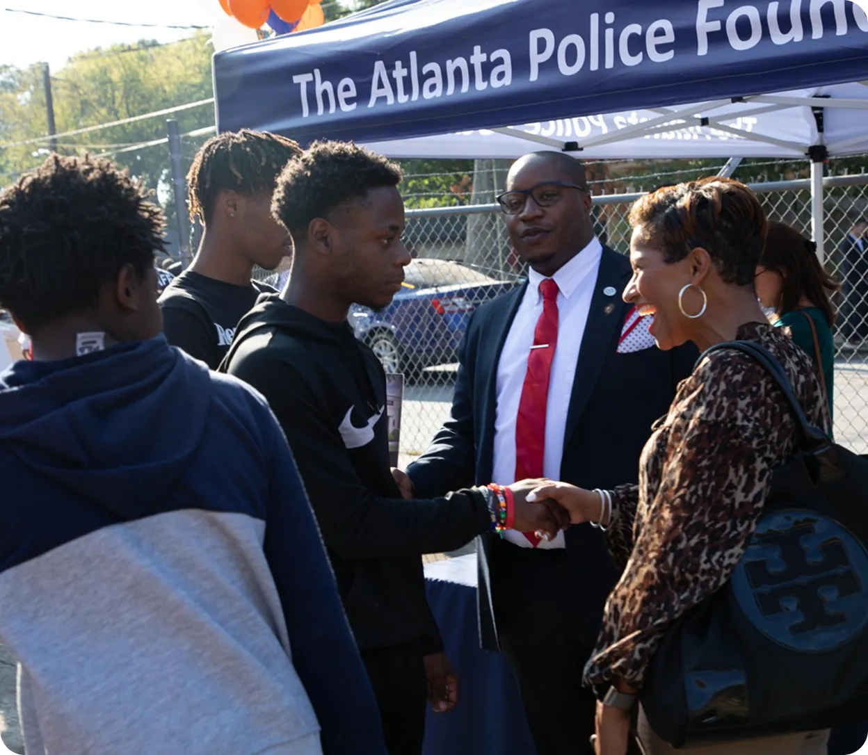 A young man shakes hands with a smiling woman near a tent labeled The Atlanta Police Foundation, with a man in a suit and other youths nearby.